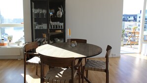 A dining area with a round table and four chairs, a bookshelf with glassware, and a view of the city through large windows. - Modern penthouse w. canal view and private rooftop terrace, 10min to city center (København)