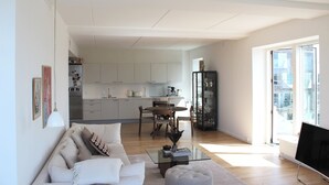 A modern living room with a sofa, coffee table, and a dining area with chairs and a table. There is a kitchen in the background with cabinets and appliances. - Modern penthouse w. canal view and private rooftop terrace, 10min to city center (København)