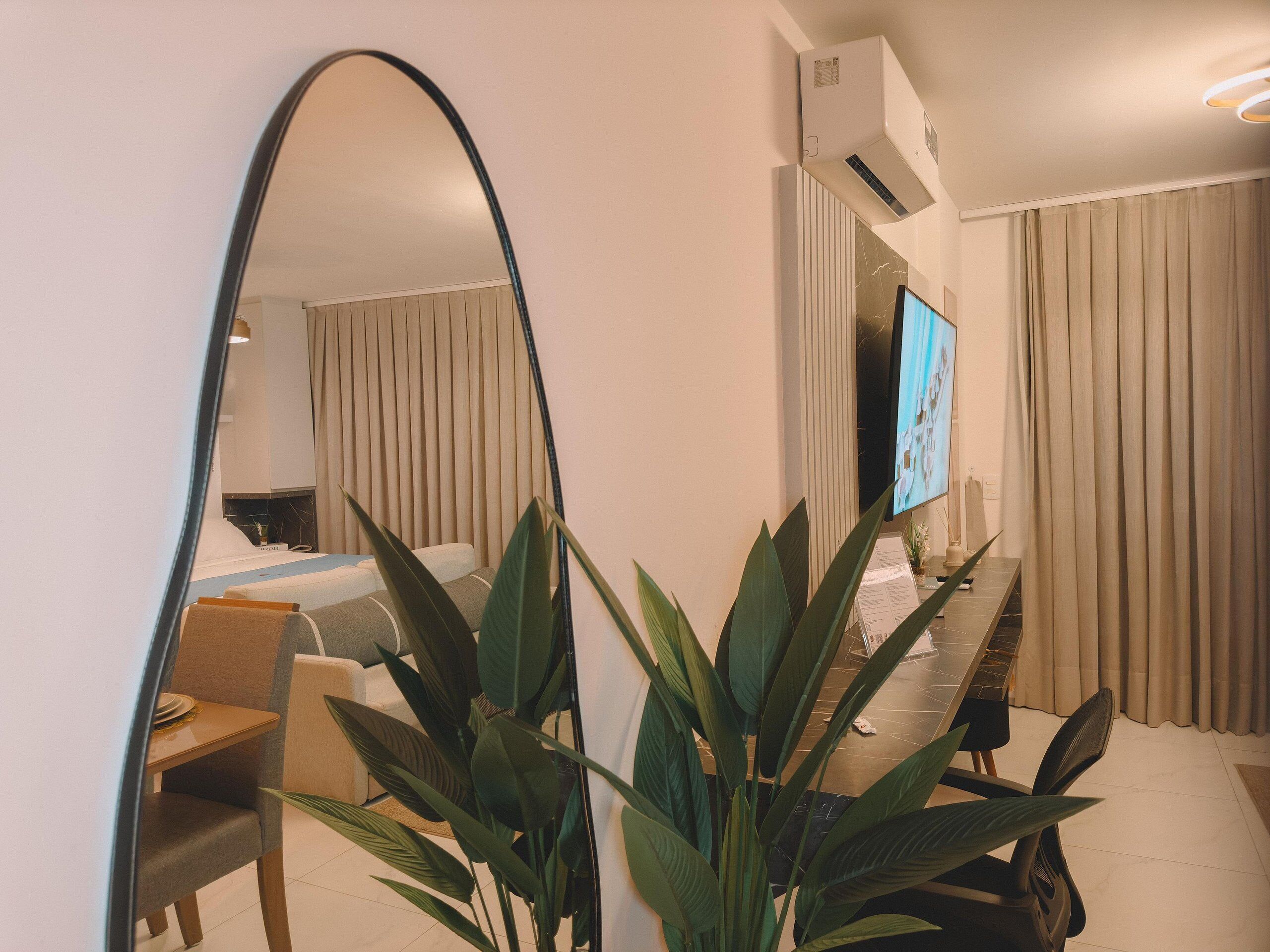 A modern hotel room with a large mirror, a flat-screen TV, a dining table, and a sofa.