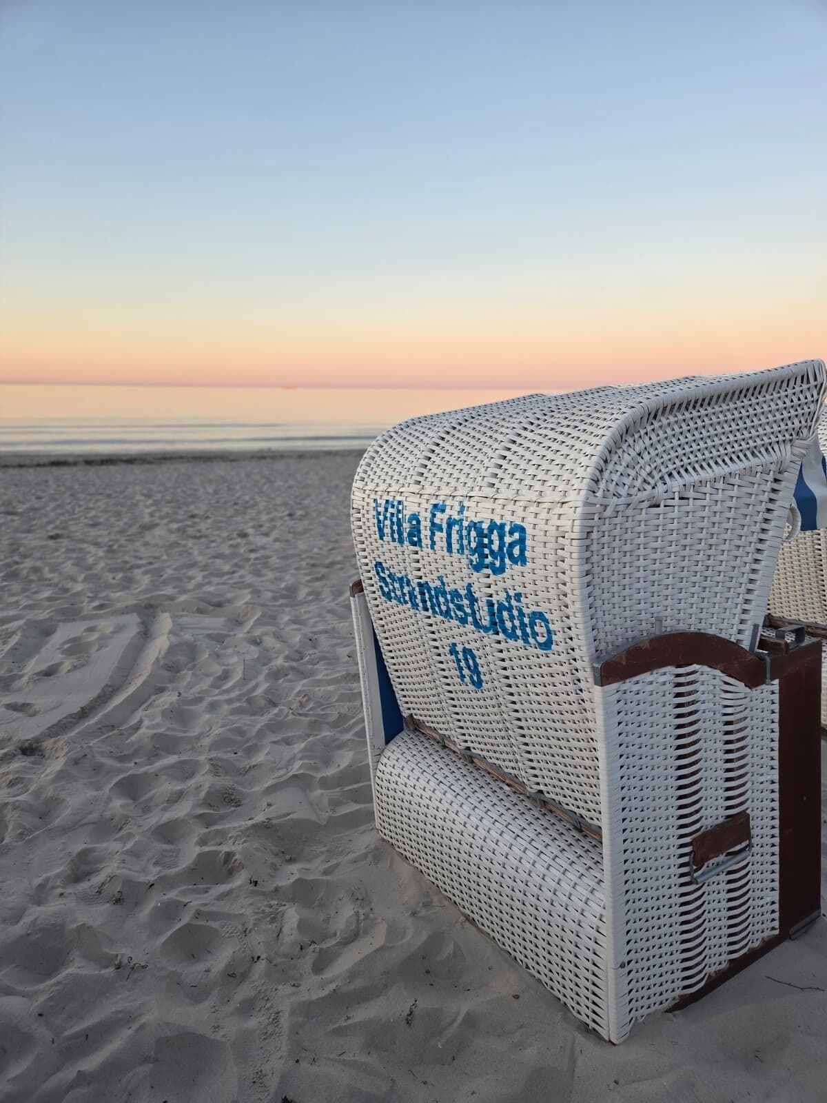 Une chaise de plage sur laquelle est inscrit « Villa Friggia Strandstudio 19 », placée sur une plage de sable.