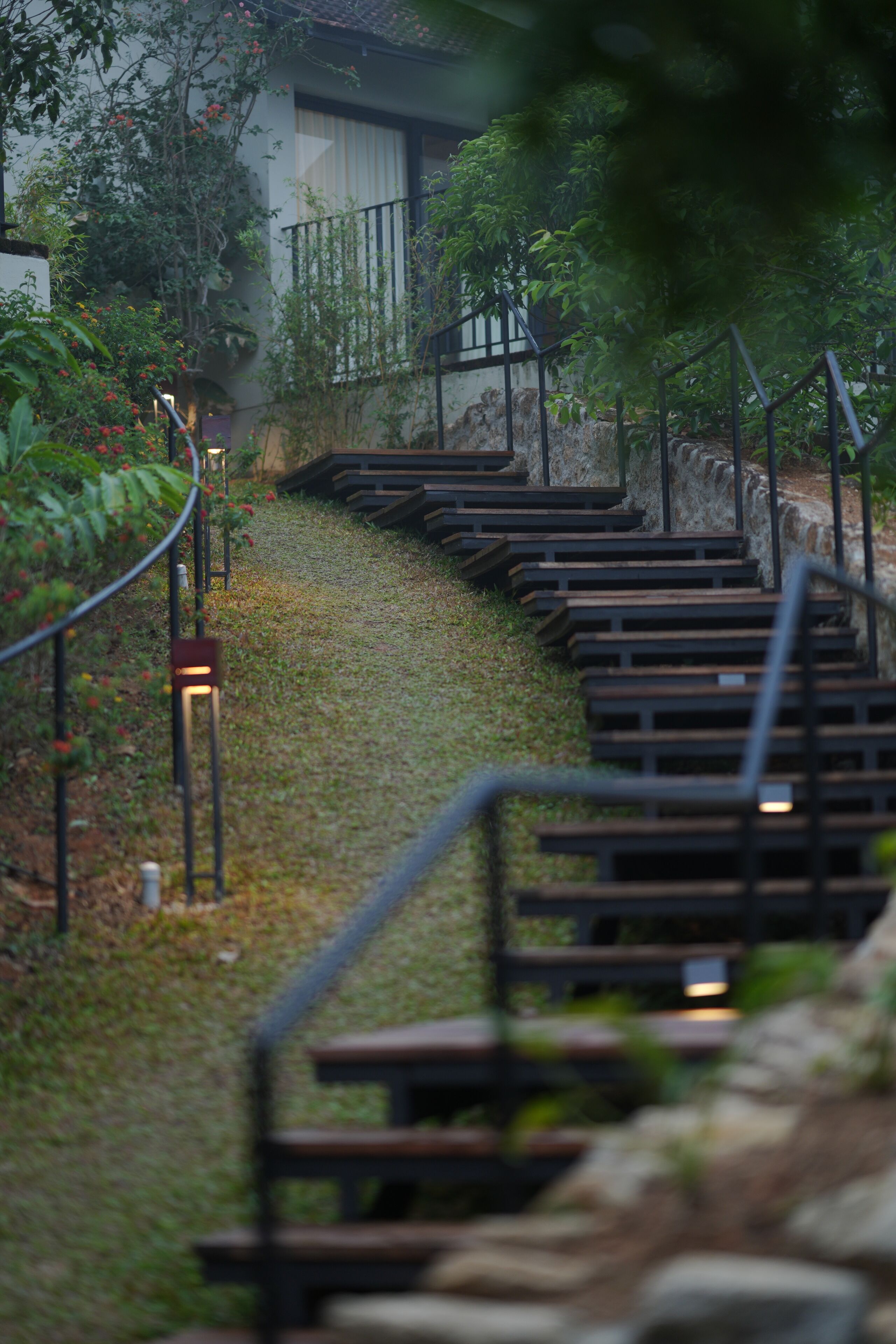 A pathway with wooden benches and metal railings, surrounded by greenery and a building in the background.