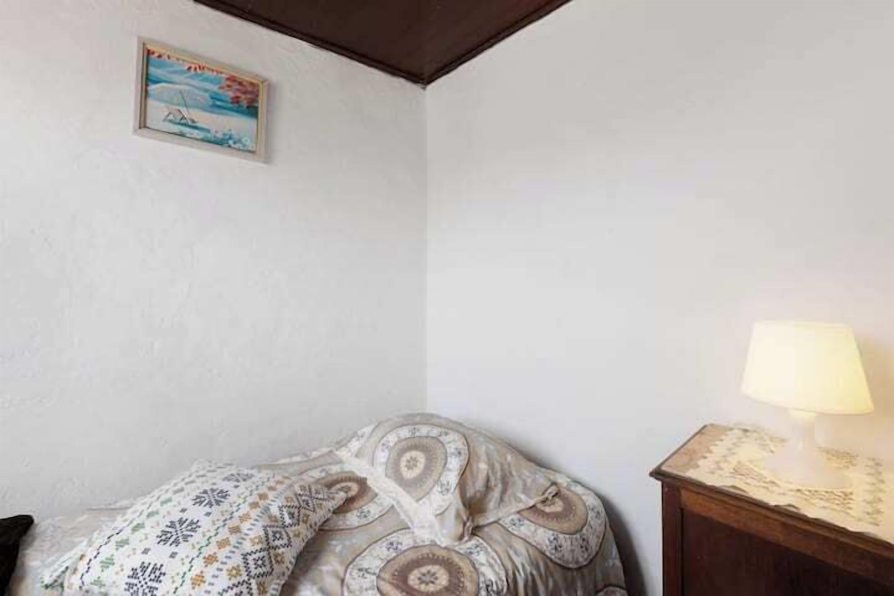 A bedroom with a bed covered in patterned bedding, a bedside table with a lamp, and a framed picture on the wall.
