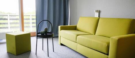 A living room with a yellow sofa, a small black table with a plant, and a window with curtains.