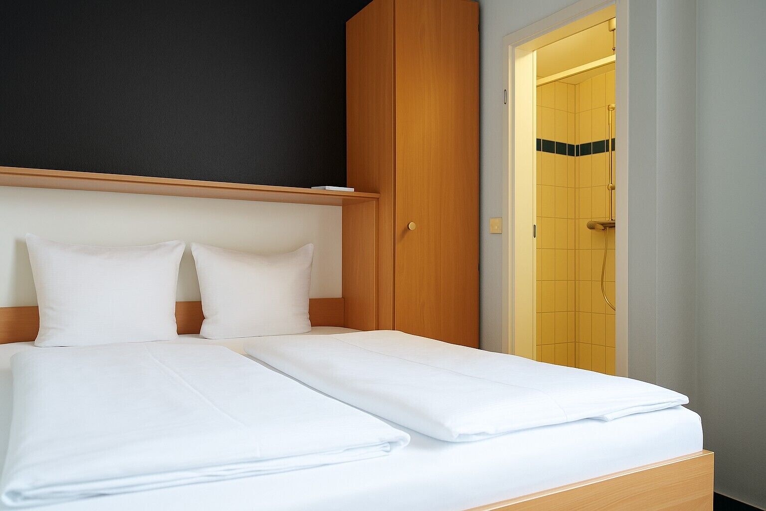 Una cama bien hecha con sábanas y almohadones blancos, un armario de madera y un baño con baldosas amarillas.