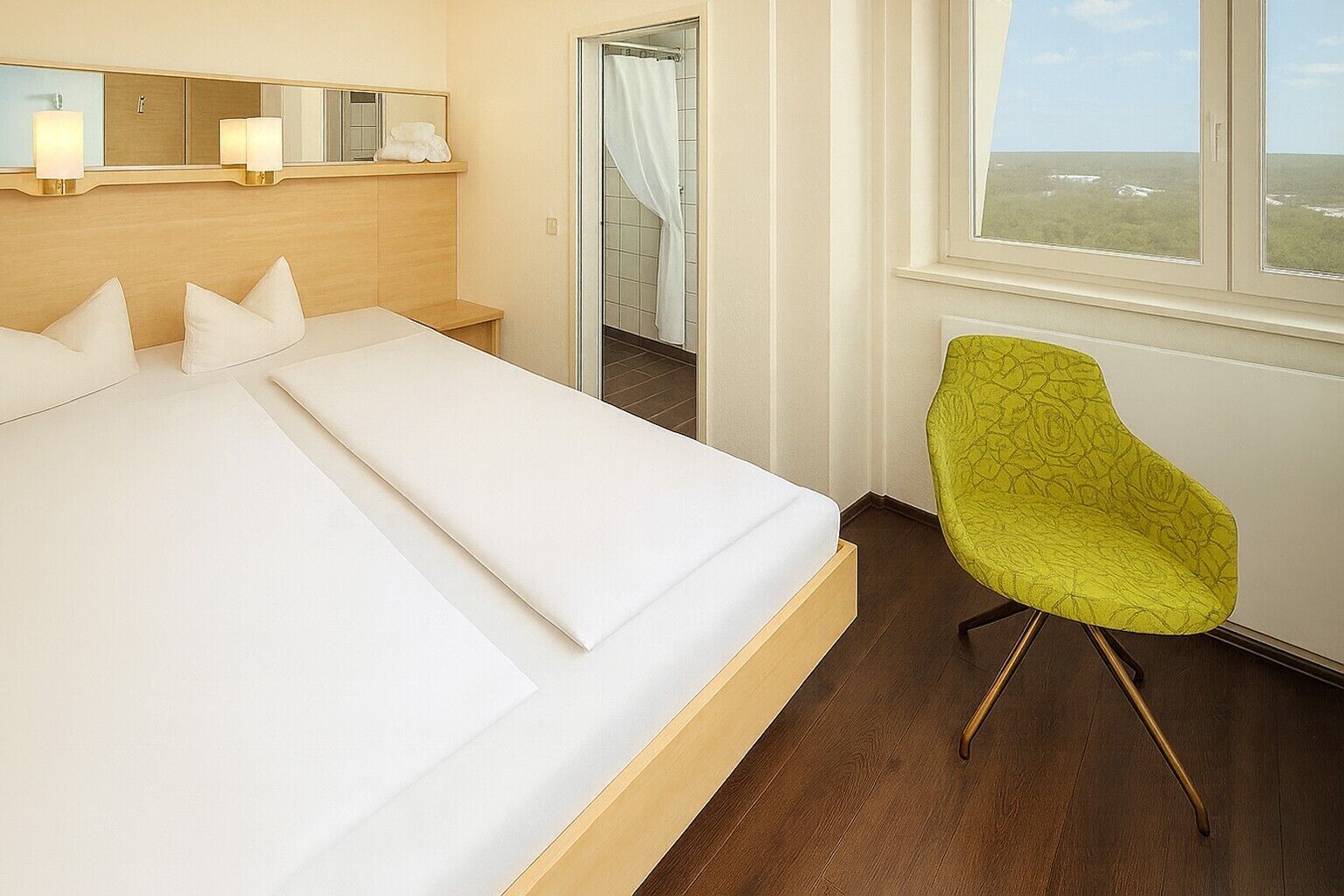 Una habitación de hotel con una cama grande, un espejo sobre la cama, una ventana con vistas y una silla verde.