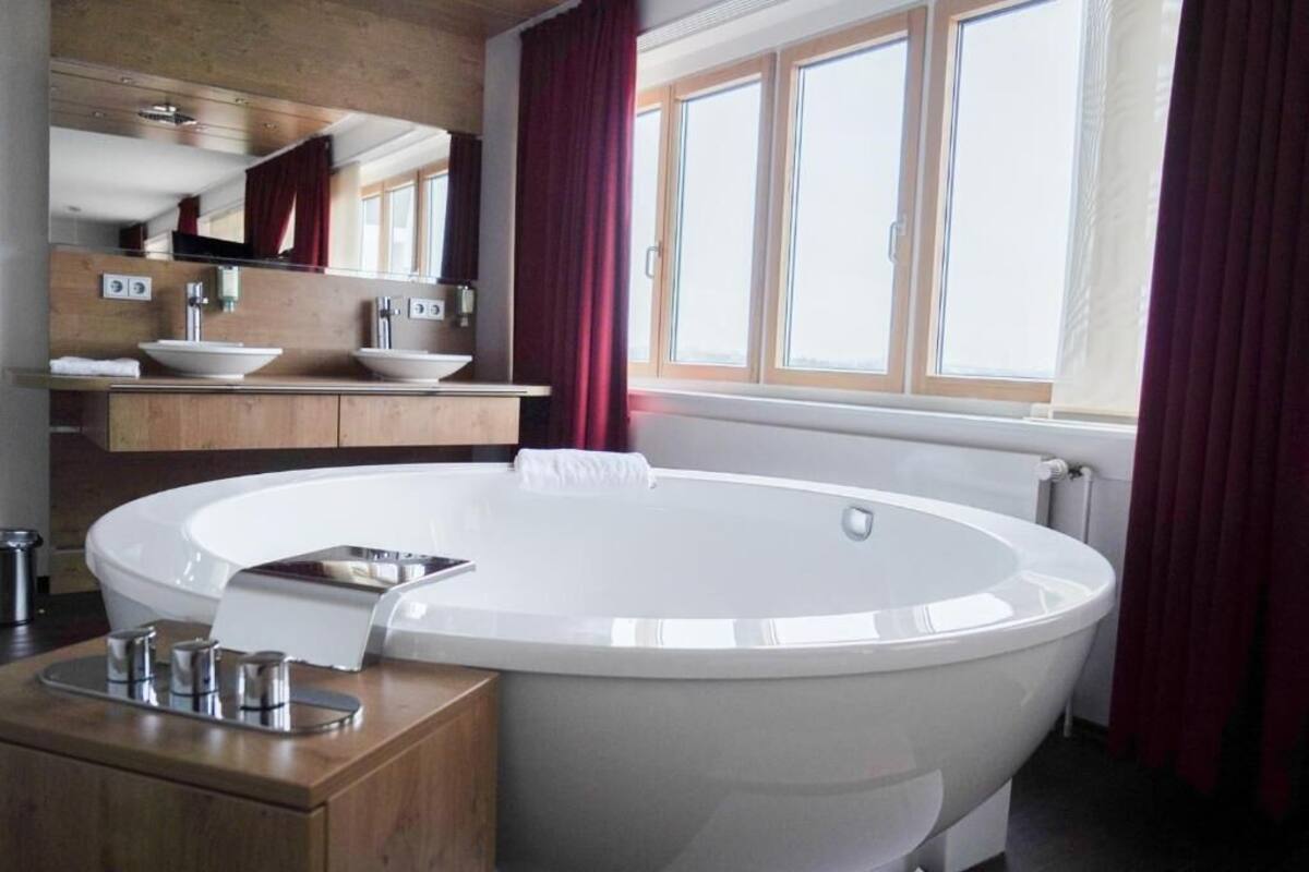 A modern bathroom with a freestanding bathtub, a wooden vanity, a large mirror, and a window with curtains.