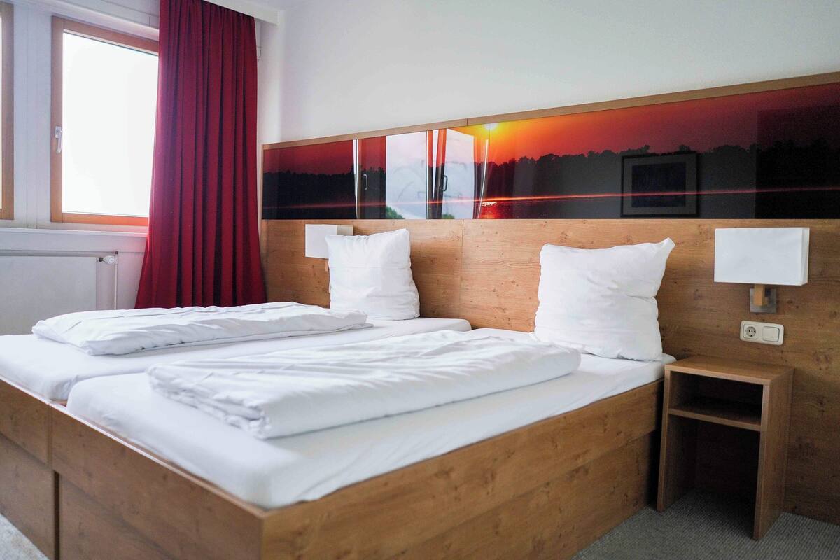 A double bed with white linens, a wooden headboard, a nightstand with a lamp, and a window with red curtains.