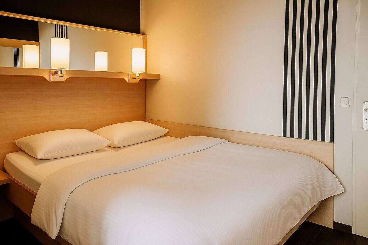 A hotel room with a bed, two pillows, a bedside table, a mirror, and a wall-mounted light fixture.