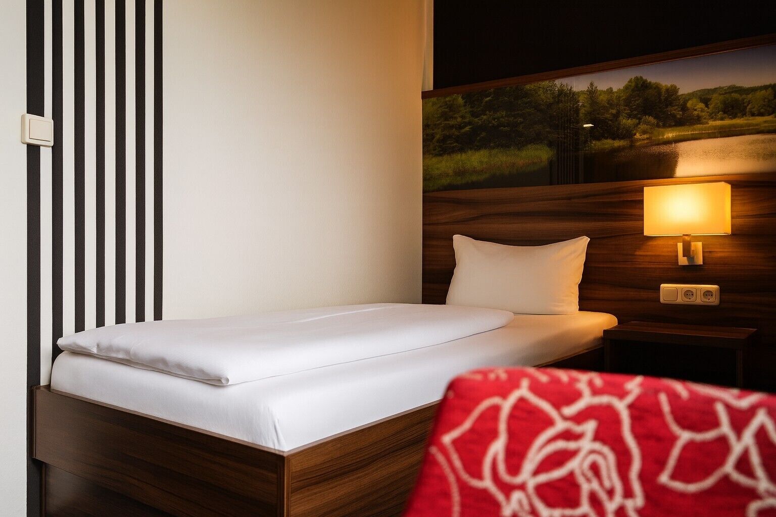 Una cama individual con colchón y almohada blancos, cabecera de madera, mesita de noche con lámpara y toma de corriente en la pared.