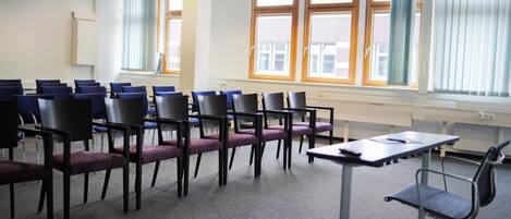 Un salón de conferencias con filas de sillas, una mesa y ventanas amplias con cortinas.