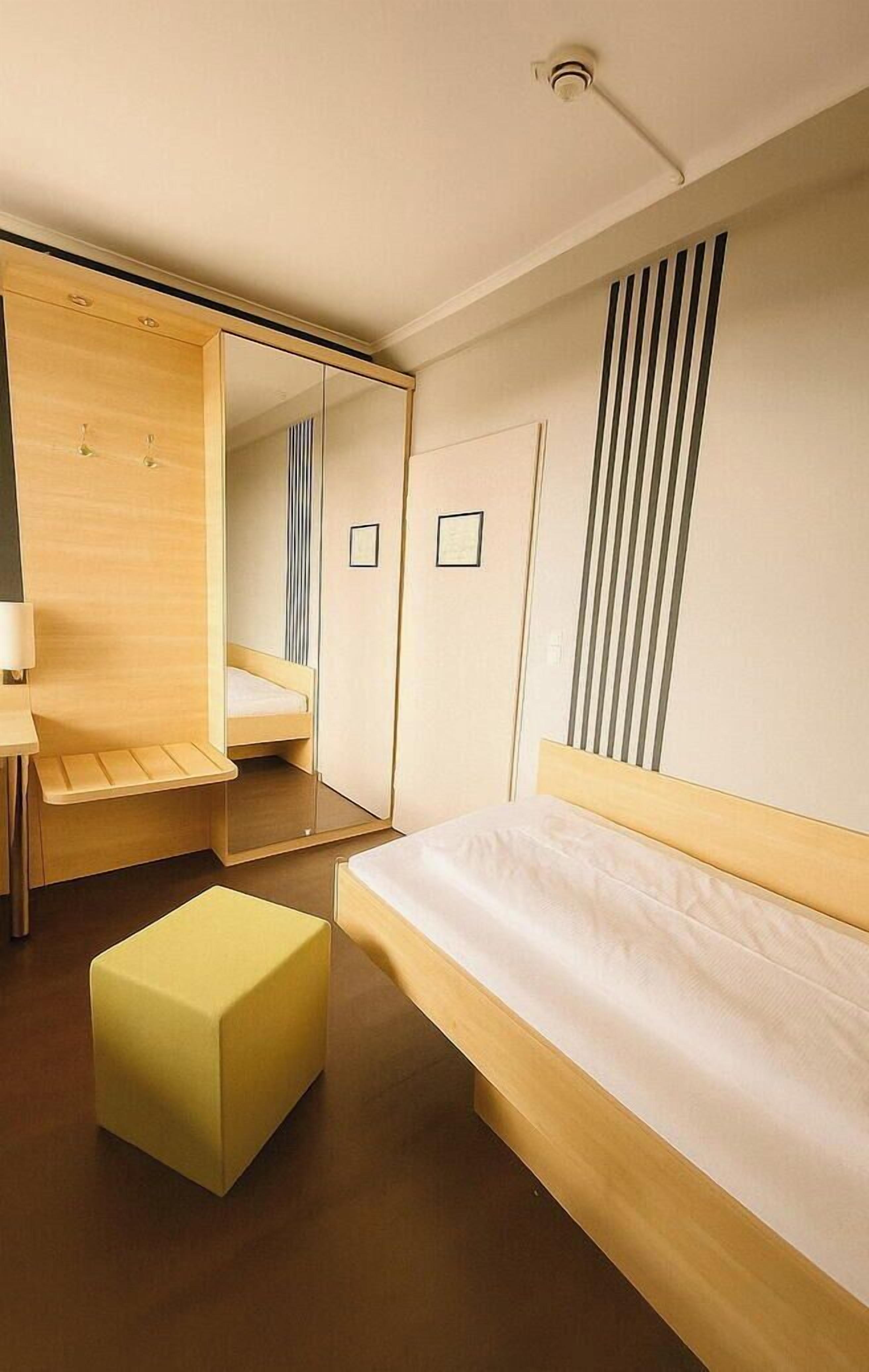 Une chambre d’hôtel moderne avec un lit en bois, un petit pouf jaune, un bureau en bois et un miroir.