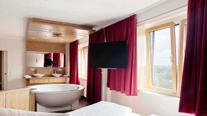Habitación de hotel con una bañera grande, un televisor montado en la pared y una cama con ropa blanca.