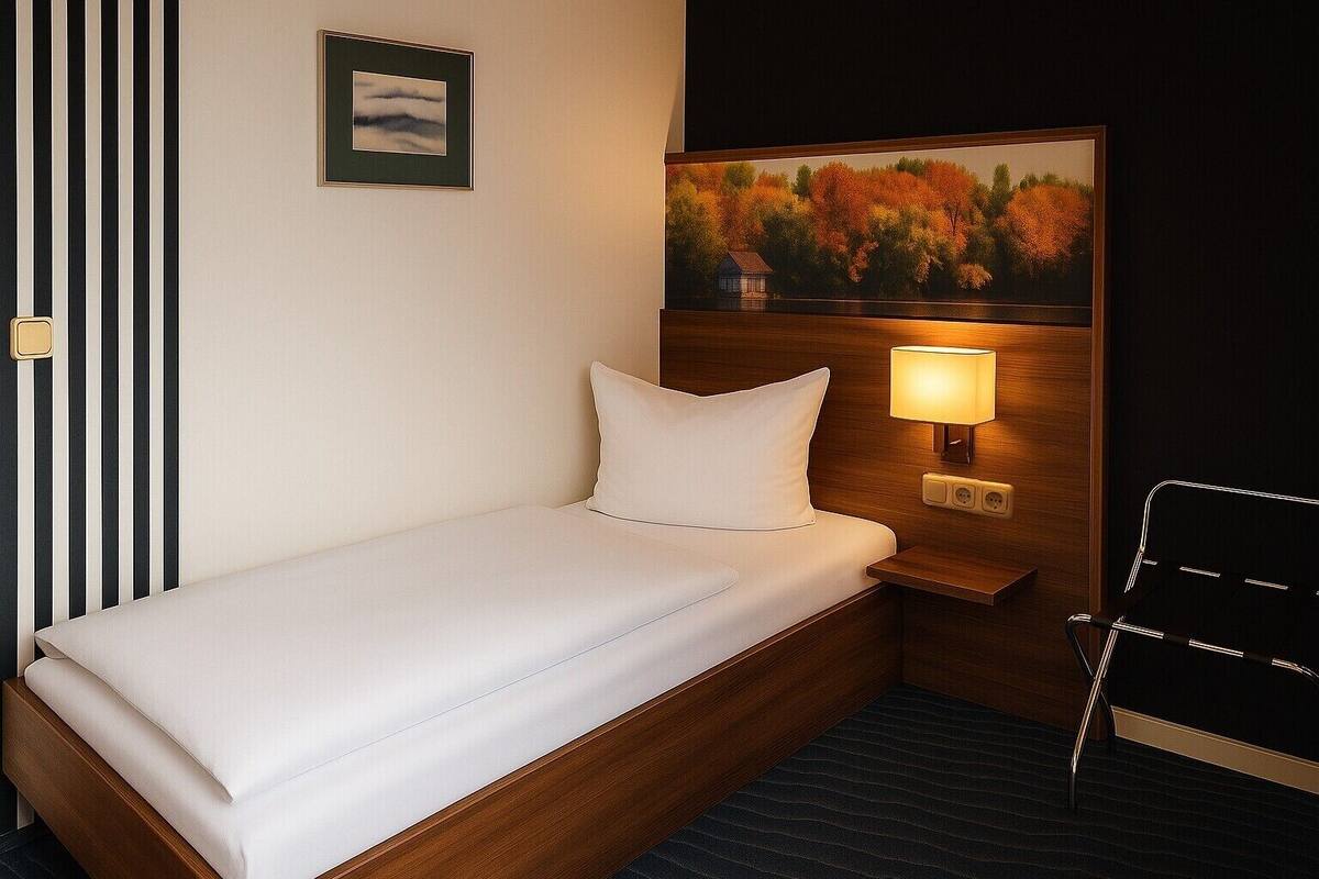 A hotel room with a single bed, a nightstand with a lamp, a framed picture on the wall, and a black chair.