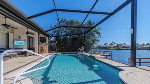 Una piscina con vistas al agua, una zona techada con asientos y un edificio con un televisor.