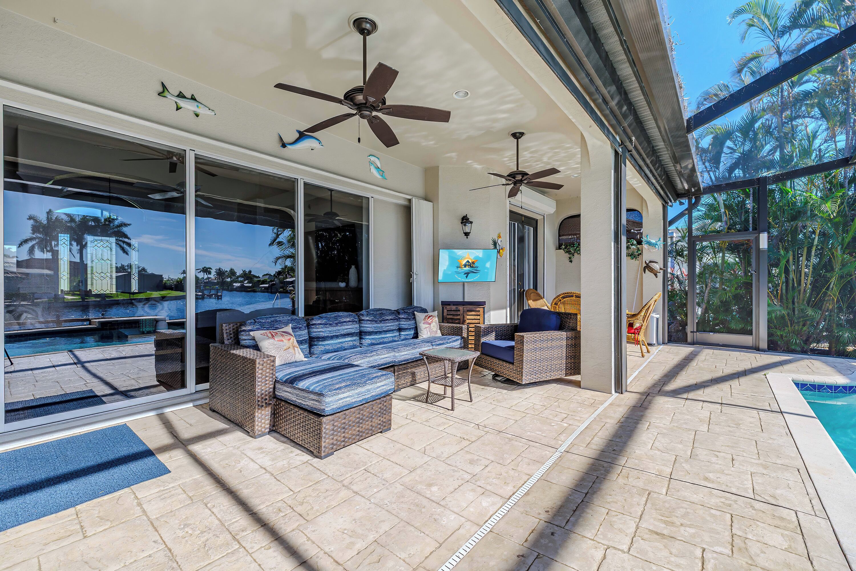 A spacious outdoor area with a pool, comfortable seating, ceiling fans, and large windows offering views of palm trees.