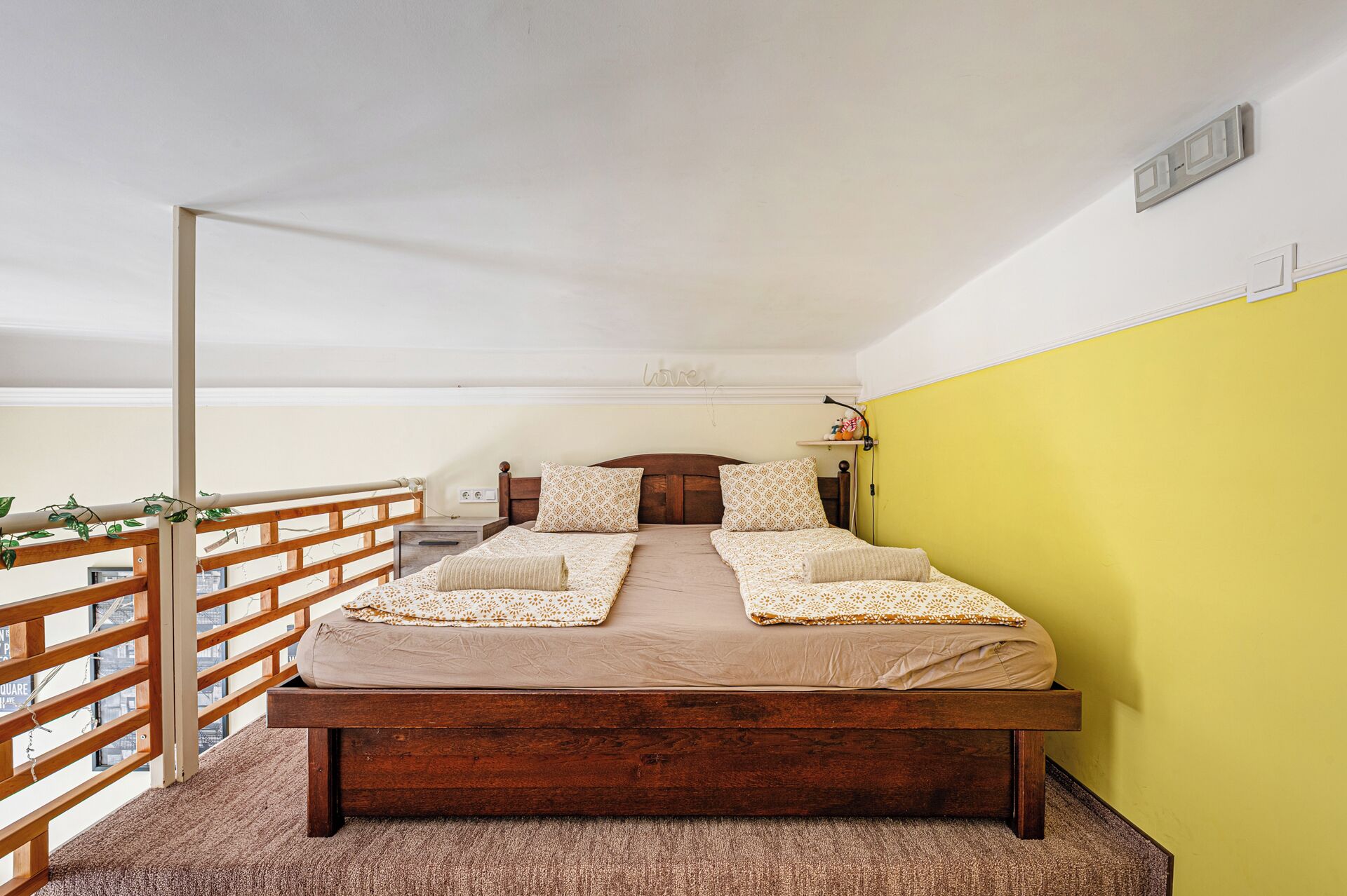 A bedroom with a wooden bed, two pillows, a nightstand, and a small shelf with decorative items.