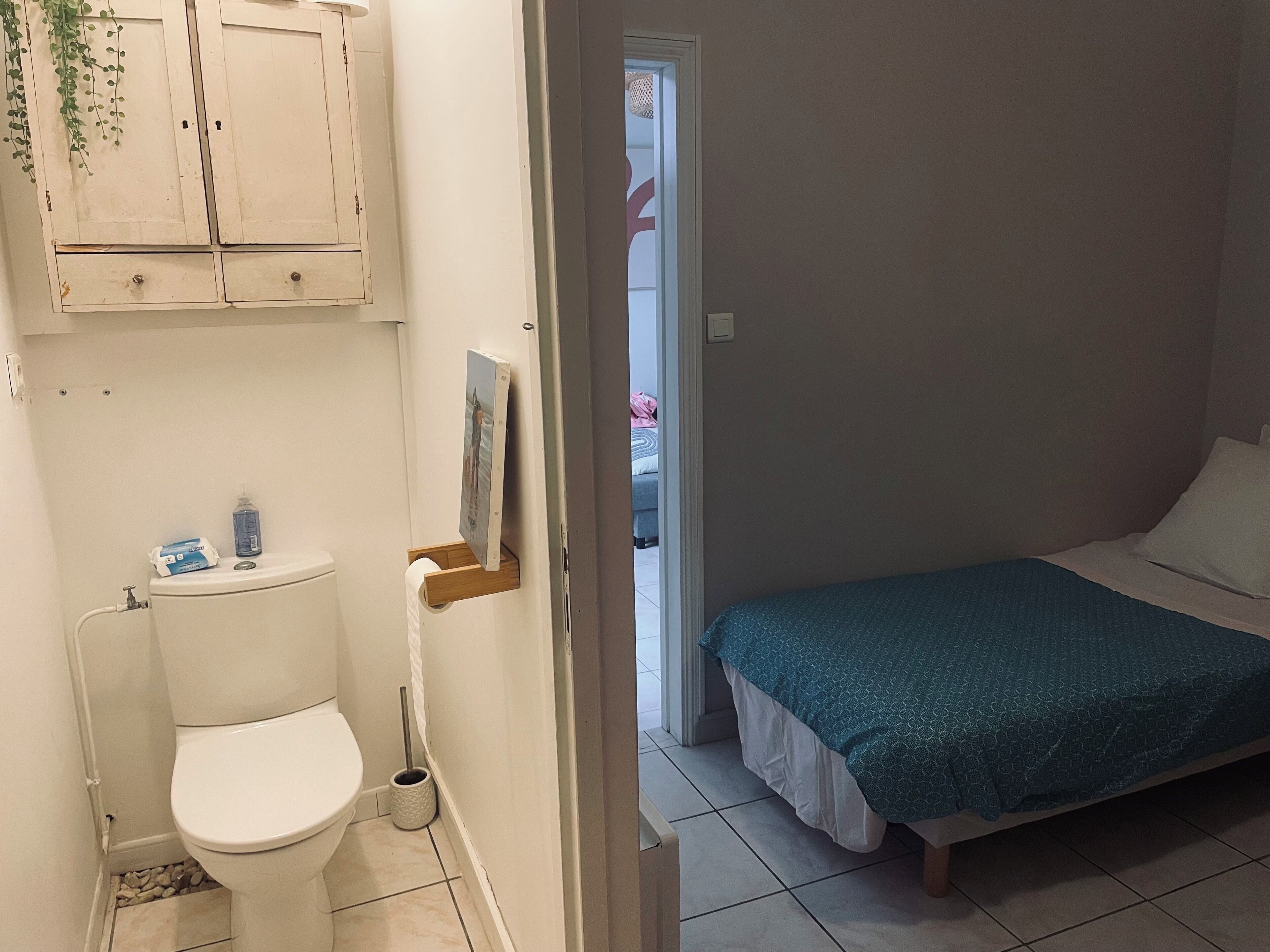 A small, clean bathroom with a toilet, a sink, and a cabinet above it. There is a bed with a blue blanket in the adjacent room.
