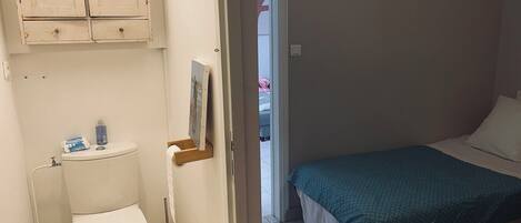 A small, clean bathroom with a toilet, a sink, and a cabinet above it. There is a bed with a blue blanket in the adjacent room.