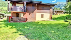 A two-story wooden house with a balcony, surrounded by a well-maintained lawn and mountains in the background.