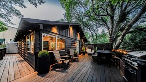 Eine Holzterrasse mit Gartenmöbeln, einem Grill und Blick auf ein Haus mit großen Fenstern und einem Baum.