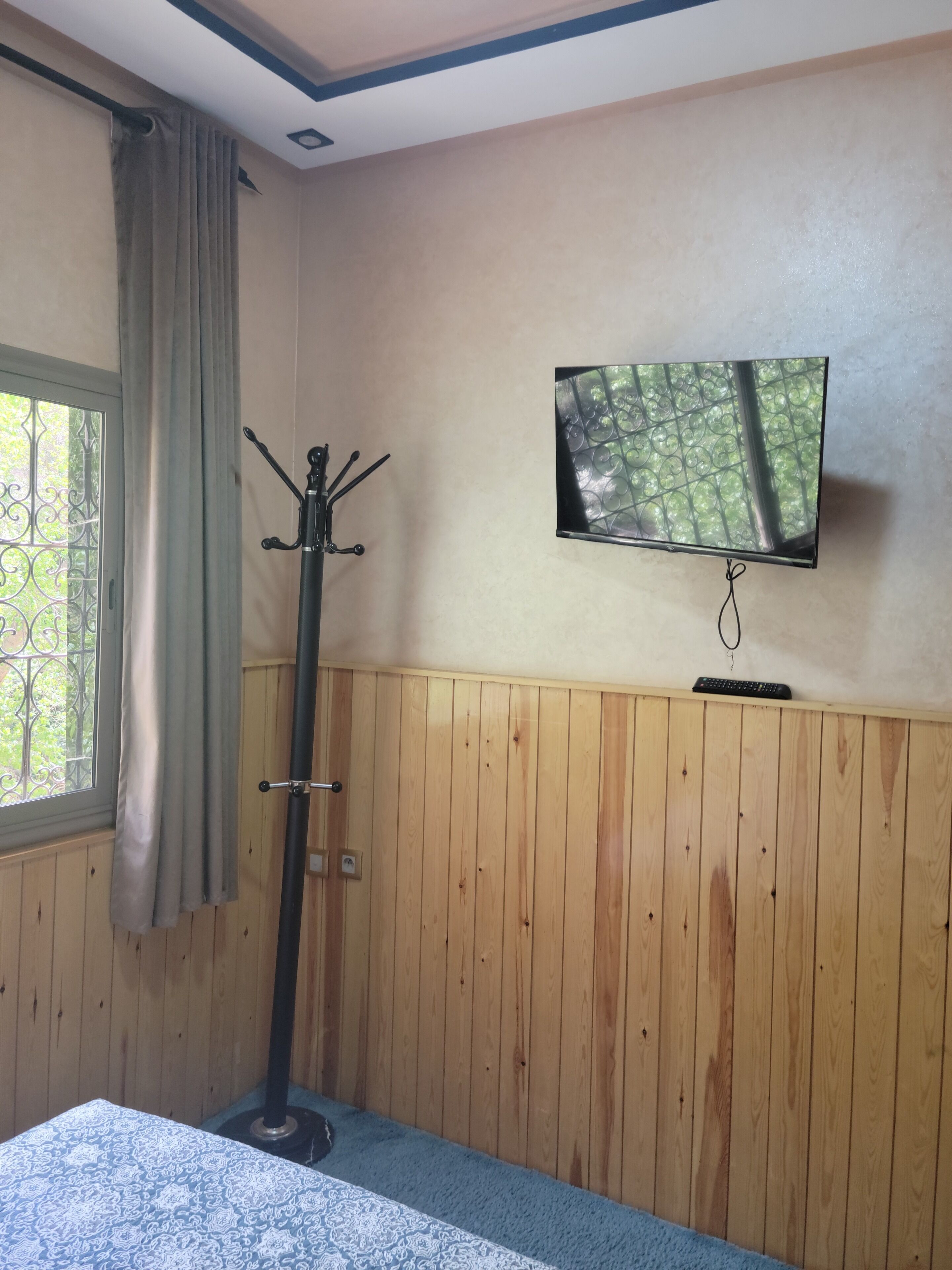 A room with wooden panel walls, a television mounted on the wall, a coat rack, and a window with curtains.