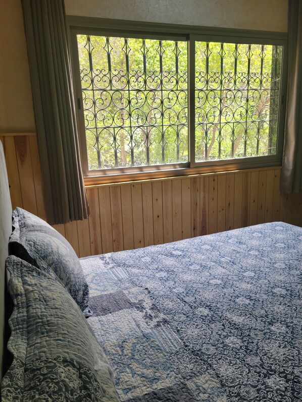 A bedroom with a wooden headboard, a large window with metal bars, and a patterned bedspread. - ATLAS OURIKA HOME (Sti Fadma)