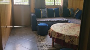 A room with a sofa, ottoman, and a small table with a floral tablecloth. There are two windows with curtains, a television on the wall, and a tiled floor. - ATLAS OURIKA HOME (Sti Fadma)