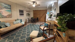 A living room with a sofa, chairs, a dining table, and a kitchen area with a TV and air conditioning unit.