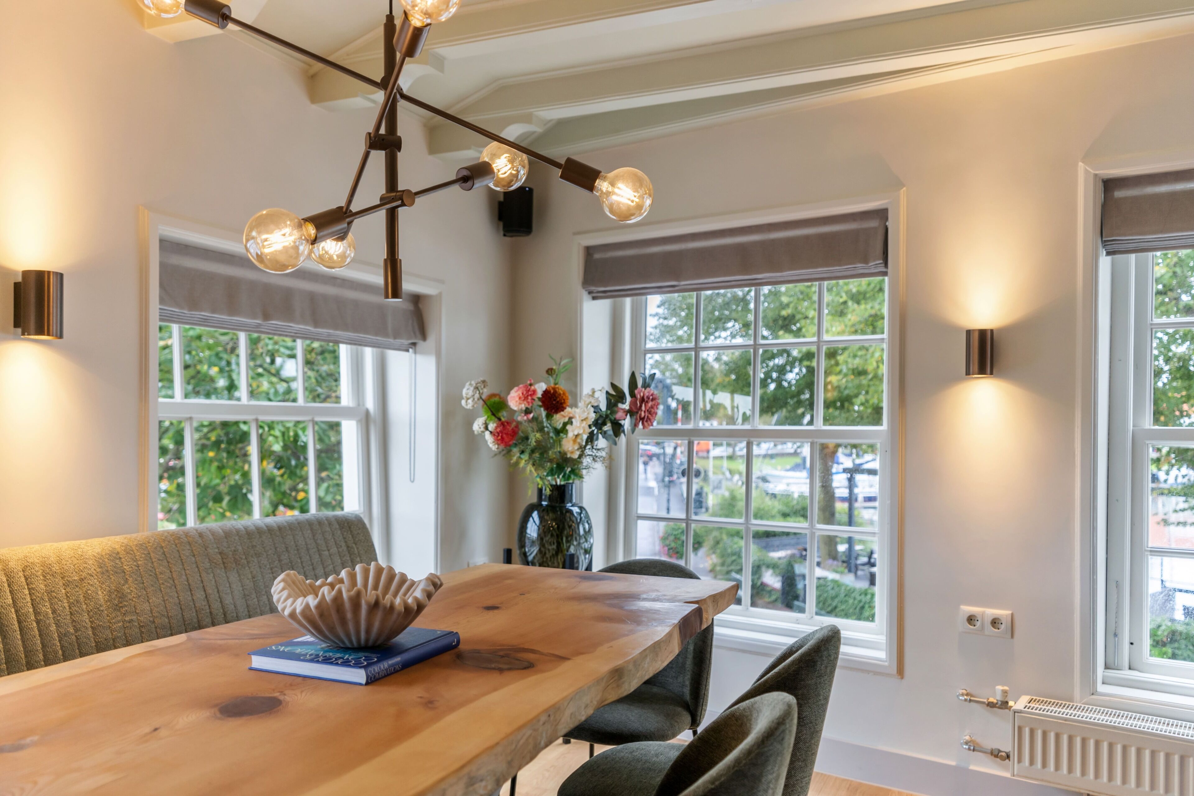 Een eetgedeelte met een houten tafel, stoelen, een vaas met bloemen en een hanglamp.