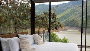 Une chambre à coucher avec un grand lit, du linge de lit blanc et une vue sur une rivière et des montagnes, visible à travers de grandes fenêtres.
