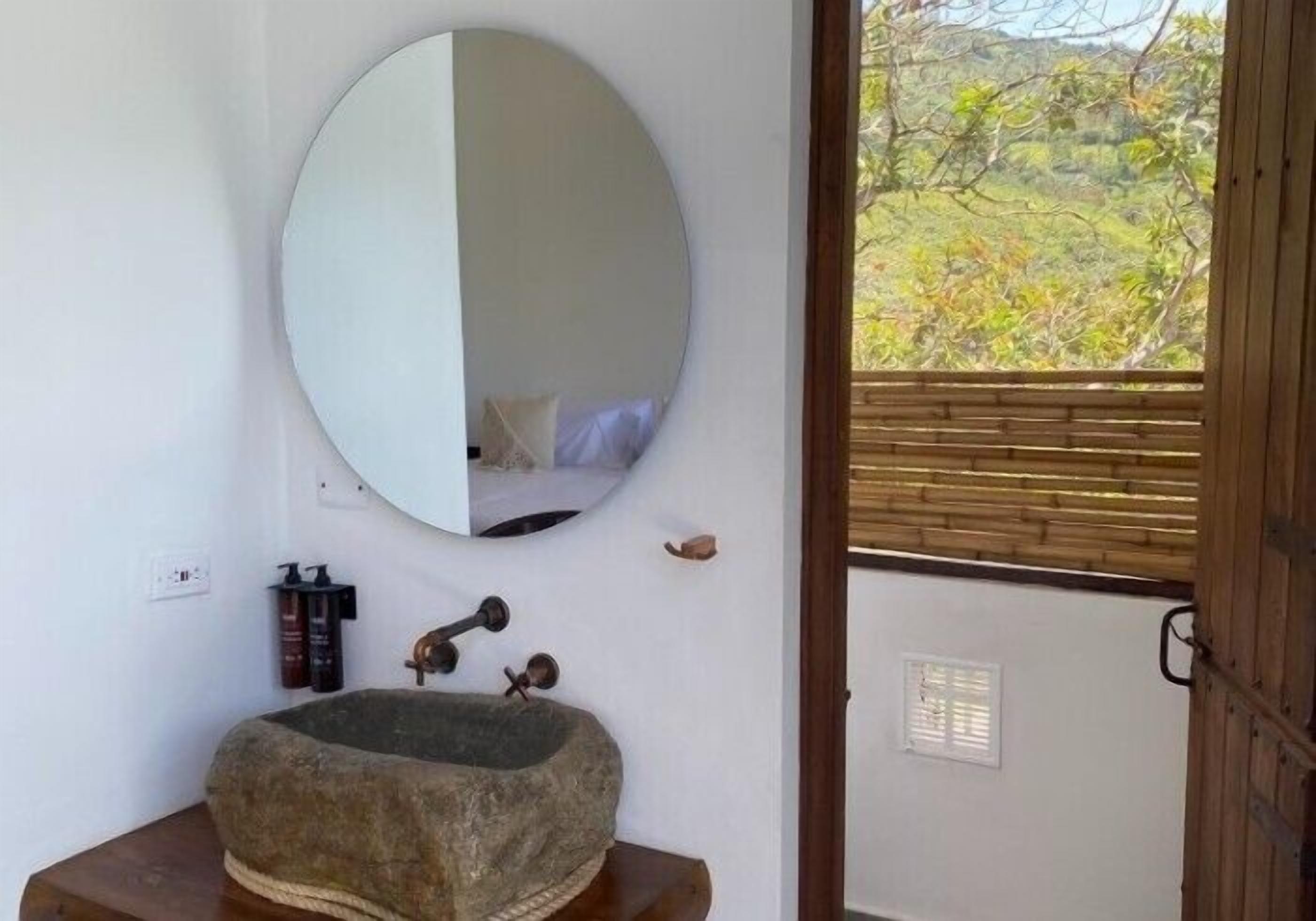 Un baño con un espejo redondo, un lavabo de piedra y una puerta de madera que da a un espacio exterior con vistas a la vegetación.