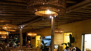 Un restaurant avec de longues tables en bois et des chaises en osier, agrémenté de lustres suspendus en osier et doté d’un sol en béton.