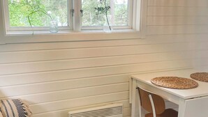Ein Zimmer mit einem weißen Tisch, zwei gewobenen Platzsets, einem Stuhl, einem Heizkörper und einem Fenster mit Blick auf Bäume.