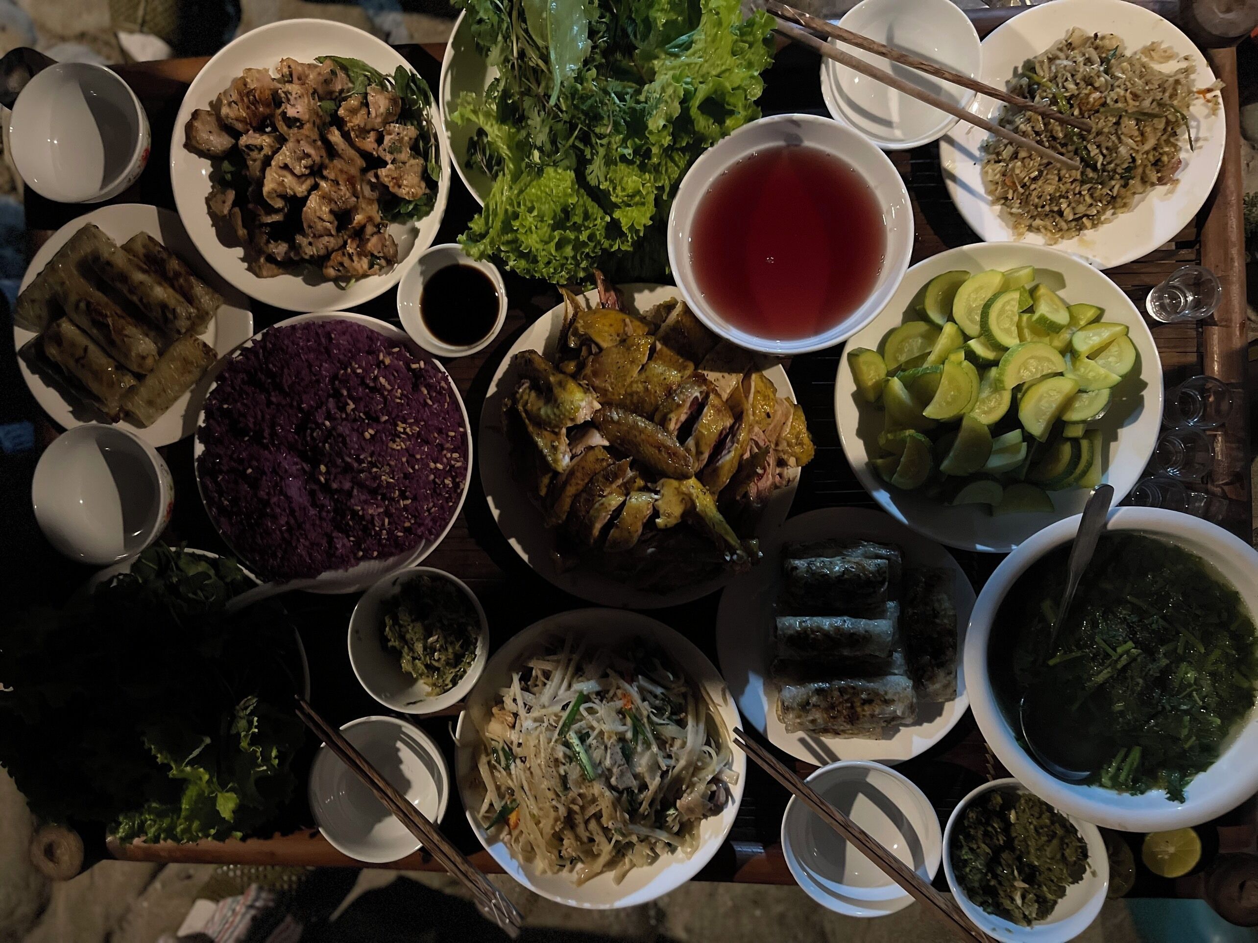 A table with various dishes, including a bowl of purple rice, a plate of grilled chicken, and several bowls of soup.