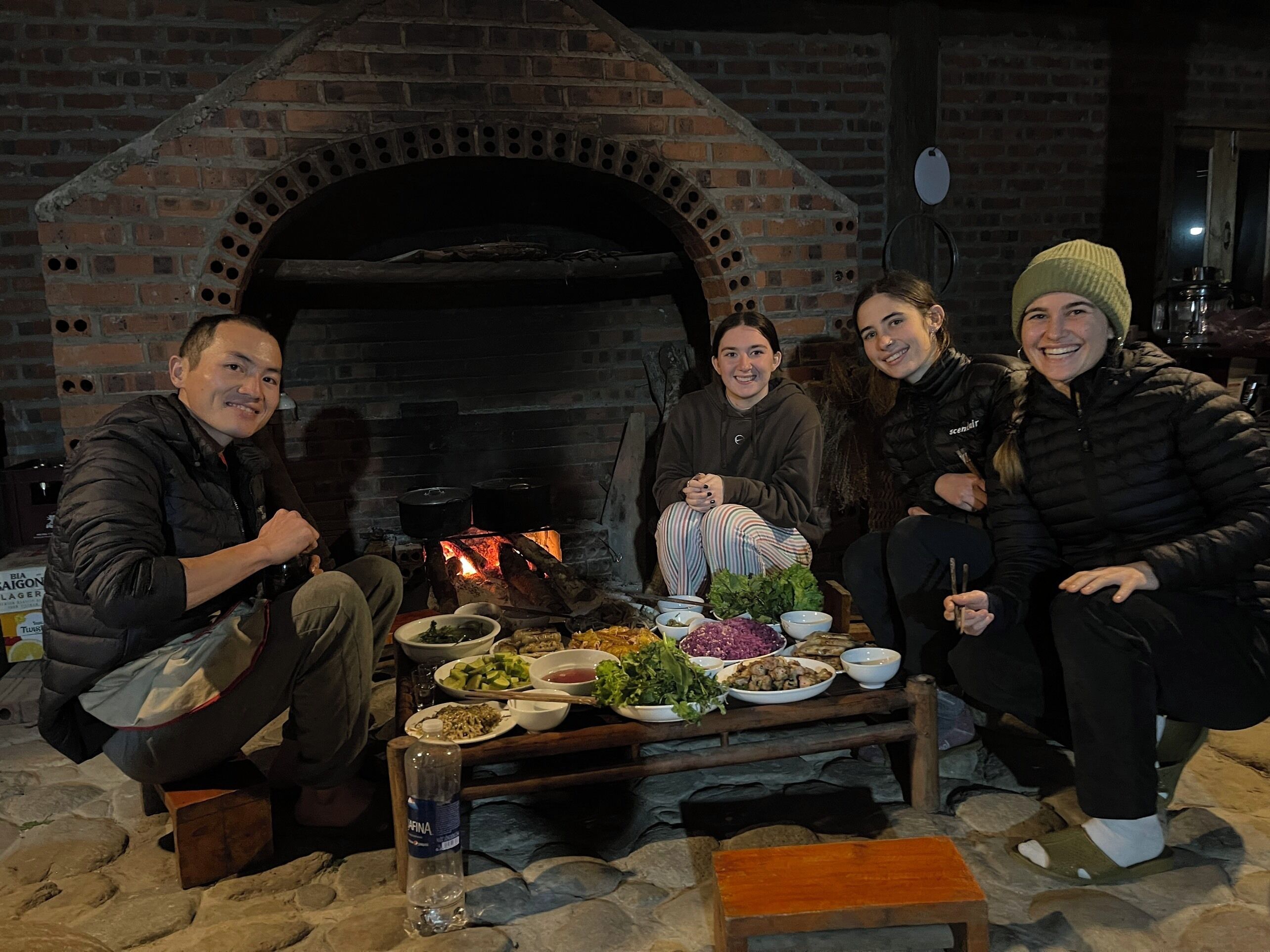A group of people seated around a table with various dishes in front of a brick oven.