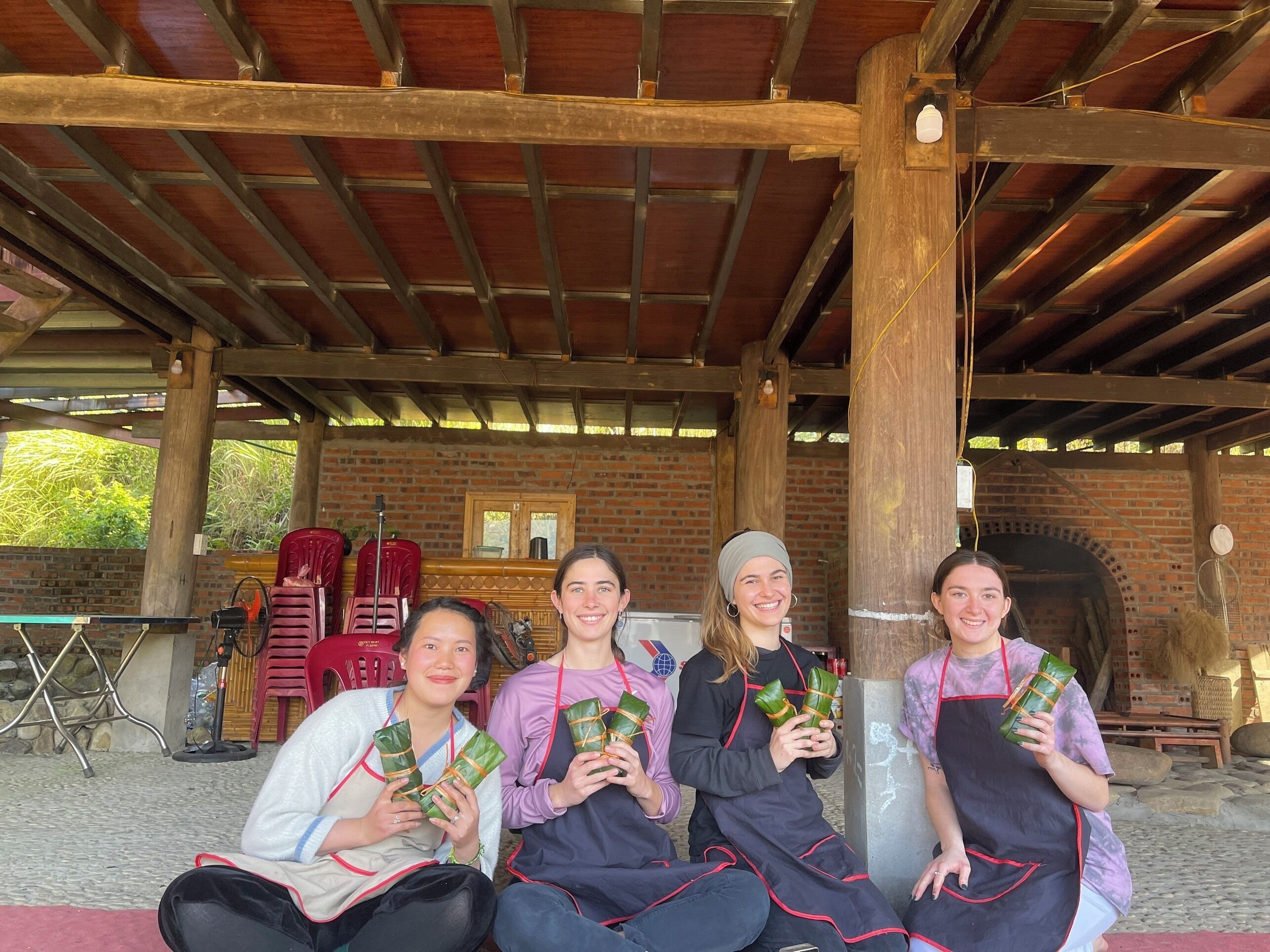 Four people seated under a wooden roofed structure with brick walls, holding wrapped food items.