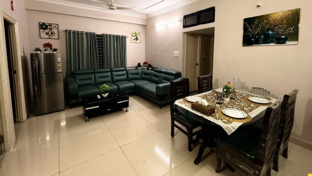A living room with a sectional sofa, a dining table set for four, a refrigerator, and a television mounted on the wall.