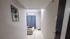 A narrow, tiled hallway leading to a bedroom with a bed, curtains, and a wall-mounted air conditioner.