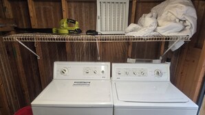 Un área de lavandería con dos lavadoras de carga frontal y un estante superior que contiene una herramienta eléctrica amarilla y un paño blanco.