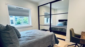Un dormitorio moderno con cama, escritorio y silla. Hay una ventana con persianas, un espejo y vistas al exterior.