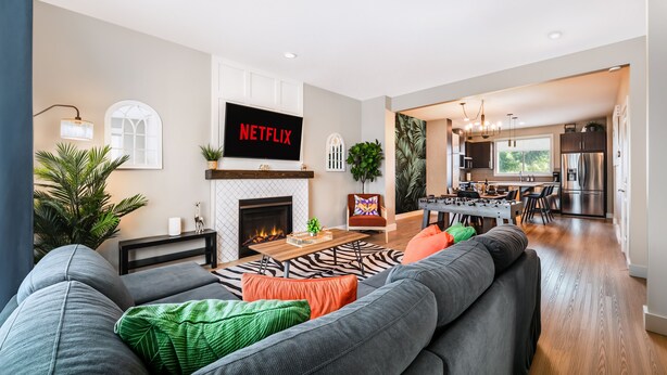 A modern living room with a fireplace, a flat-screen TV displaying 'NETFLIX', and a dining area with a kitchen in the background. - Jungle themed house for groups up to 13 (Edmonton)