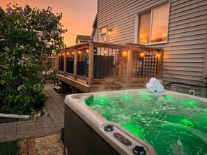A hot tub with a wooden deck and string lights, surrounded by a garden and a house.