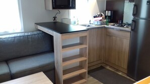A compact kitchen with a built-in microwave, a sink, and a stove. There is a wooden cabinet with open shelves and a grey sofa in the living area.