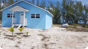 Een laag huis met een blauwe gevel en een witte veranda, met een kleine voortuin en een witte auto die er geparkeerd staat.