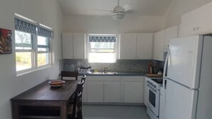 Een keuken met witte kastjes, een fornuis, een koelkast, een eettafel met stoelen en een raam met gordijnen met dessin.