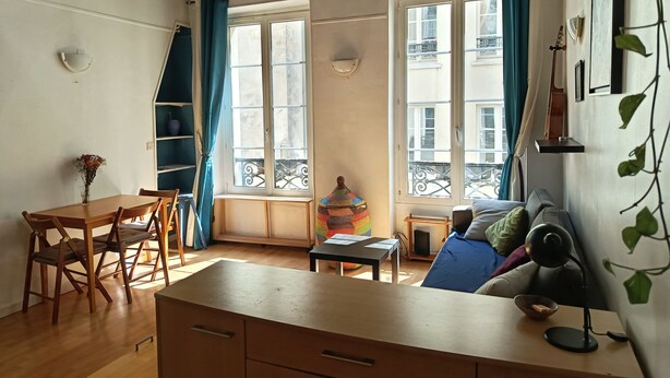 A cozy room with a wooden table, chairs, a sofa, and a desk with a lamp. There are two windows with curtains, a plant, and a shelf with items. - Cosy studio in the heart of Paris (Paris)