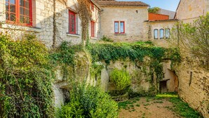A stone building with red window frames, a small garden area, and a stone wall with vegetation. - Maison Azur - Pool - Private cinema - 15 min Saumur (Doué-en-Anjou)