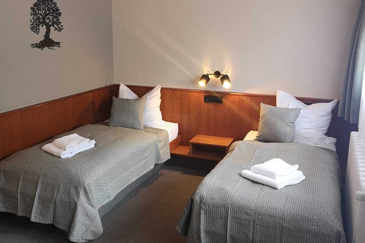 A hotel room with two single beds, a nightstand, a wall-mounted lamp, and a tree decal on the wall.
