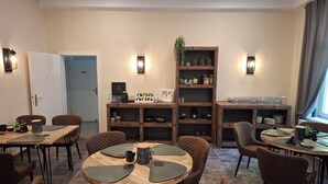 A modern dining area with round tables and cushioned chairs, a wooden bookshelf with various items, and wall-mounted lighting.