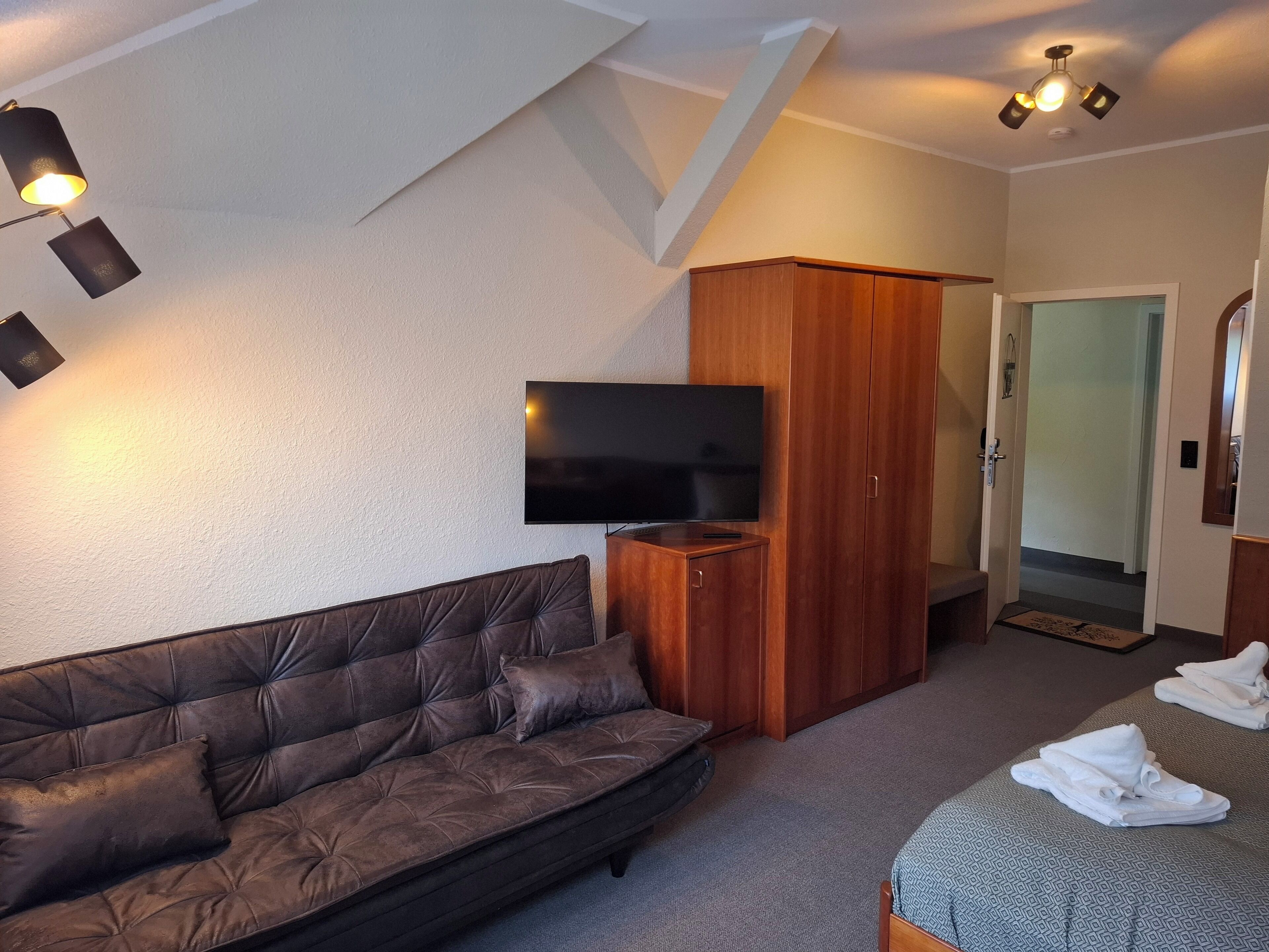 Ein modernes Zimmer mit braunem Ledersofa, einem Holzschrank, einem Flachbildfernseher und einem Bett mit Handtüchern.